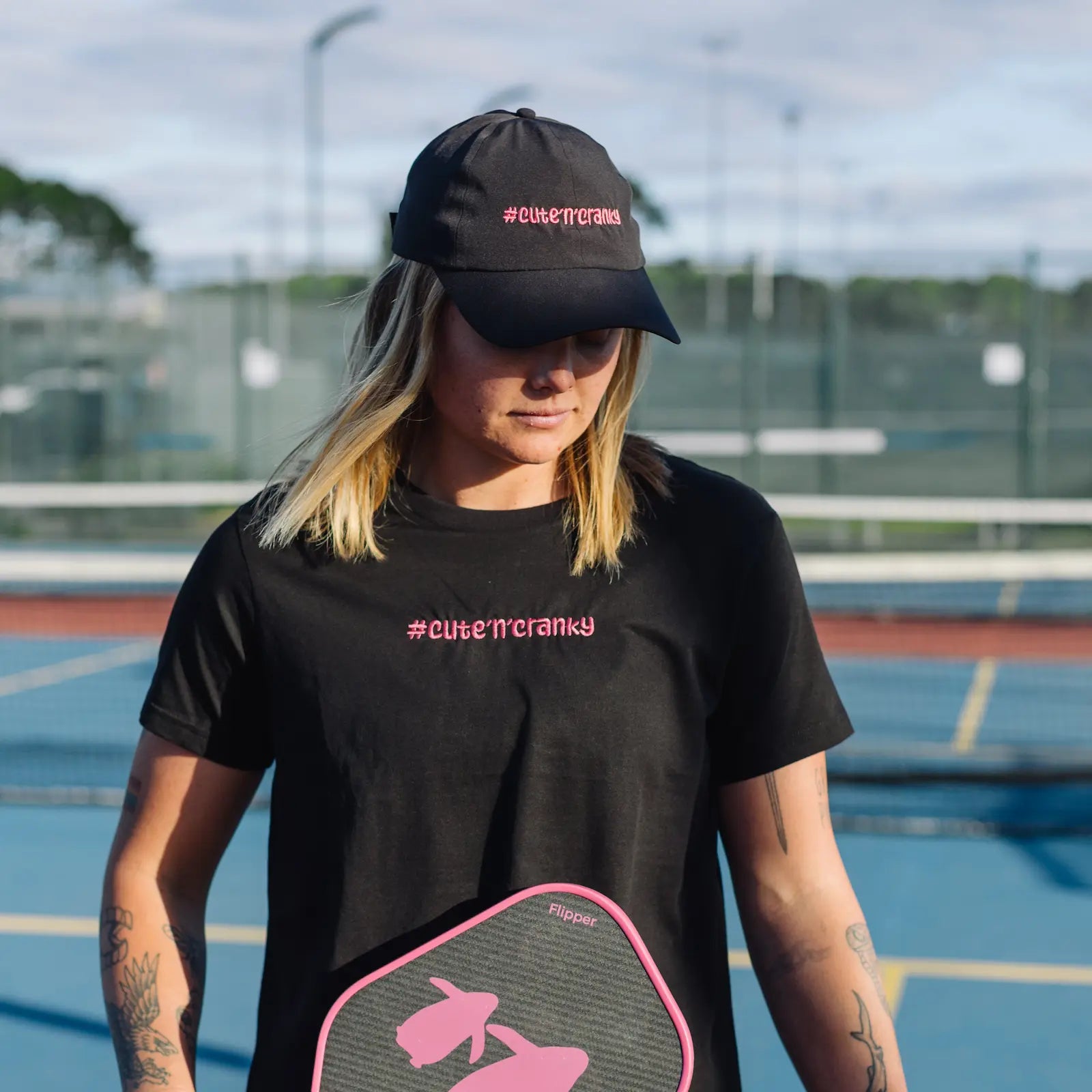 Woman on a pickleball court wearing a black t-shirt and cap with '#cute'n'cranky' text, holding a pink and black pickleball paddle. | Angry Penguins
