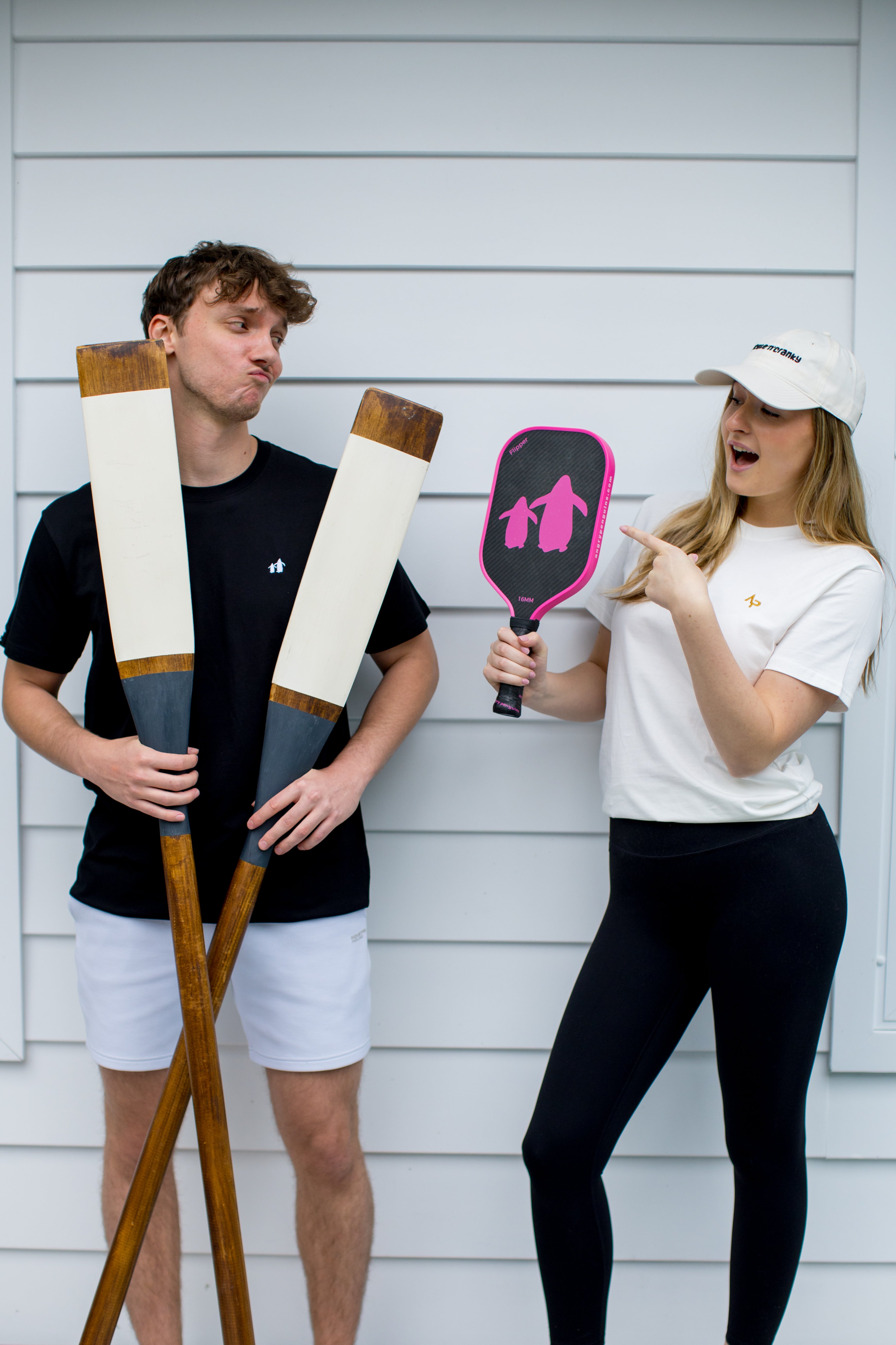 Two people holding paddle sports equipment against a white wall wearing Angry Penguins tees and cap