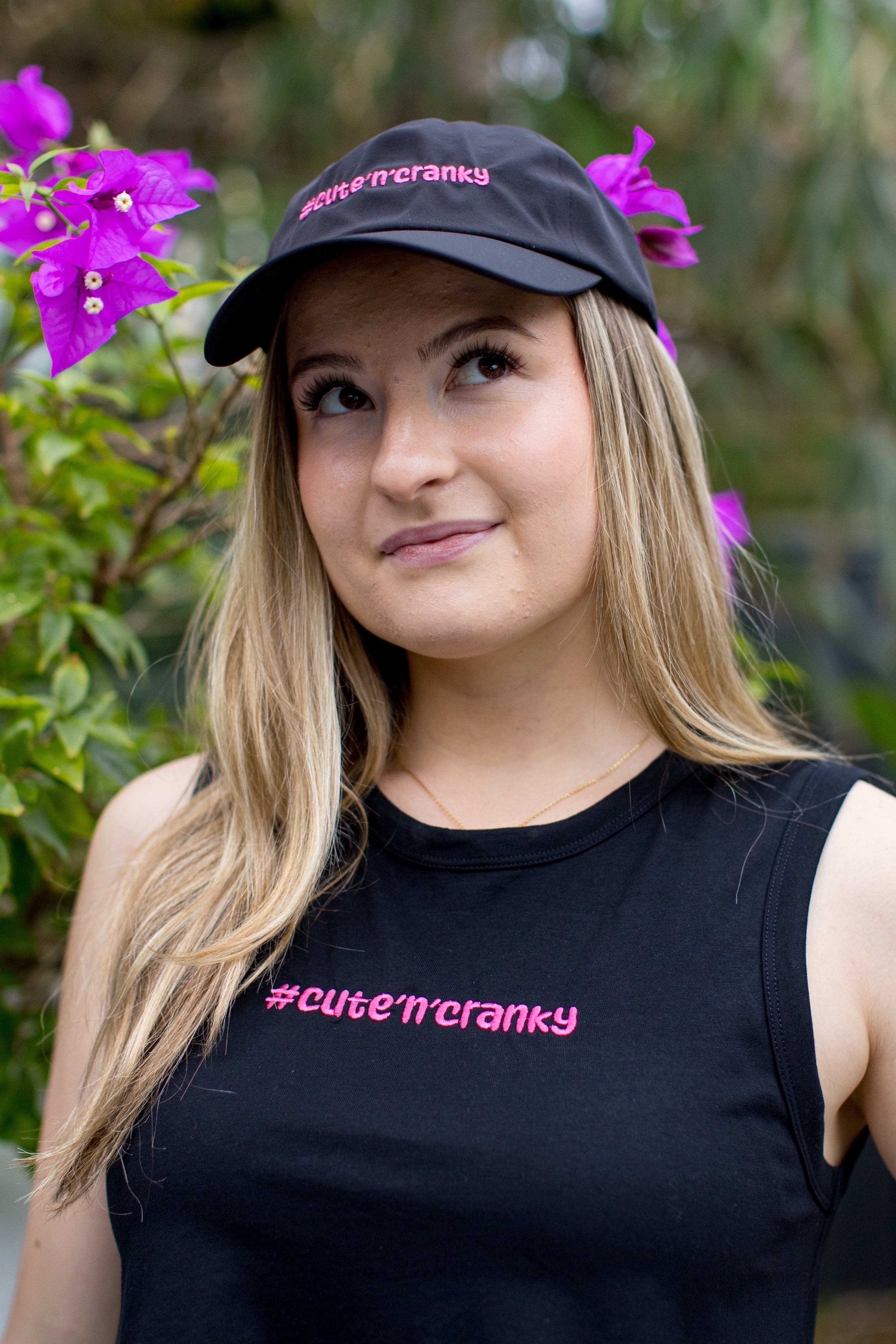 Woman wearing a black cap and tank top with '#cute'n'cranky' text, surrounded by purple flowers. | Angry Penguins