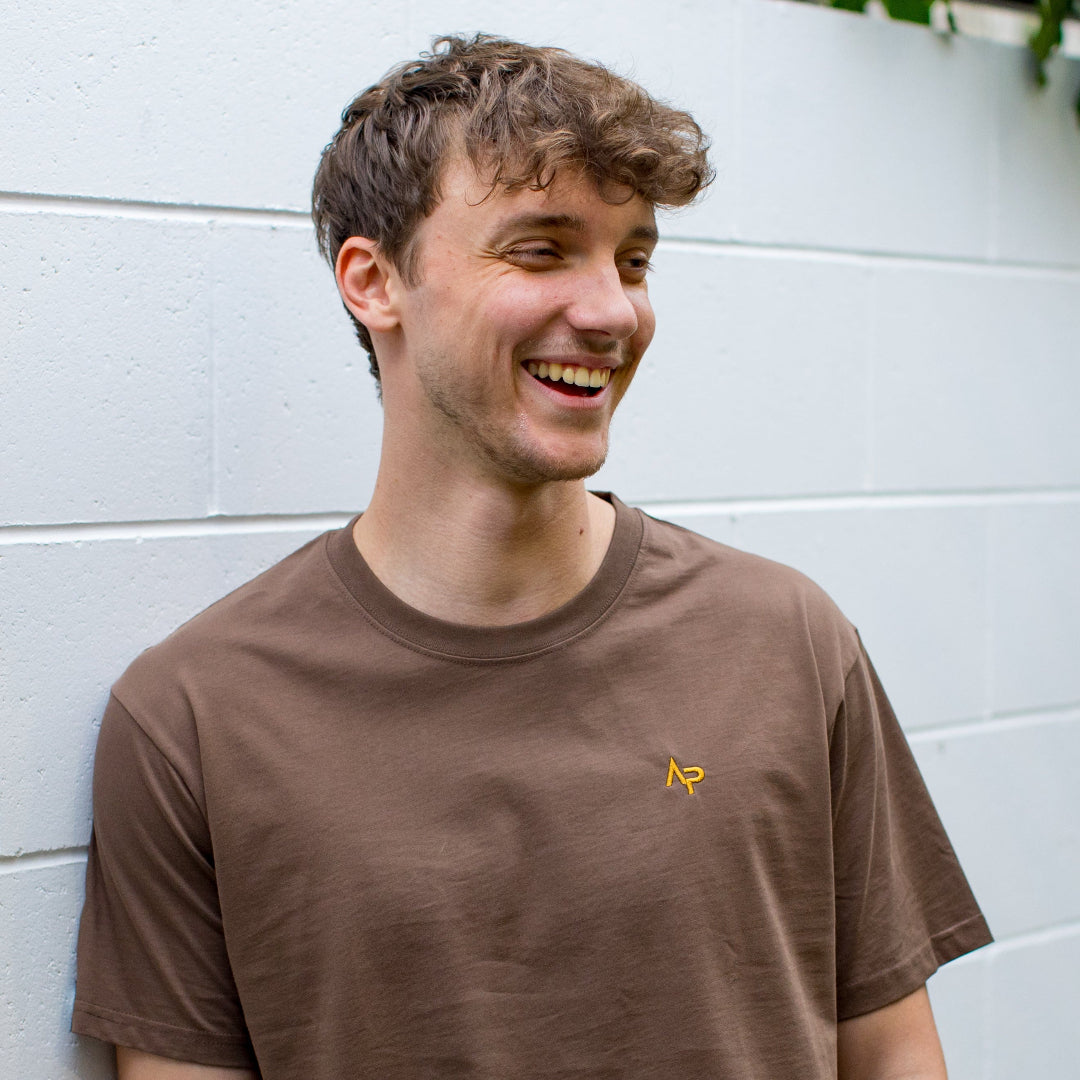 Man wearing a brown t-shirt with a logo, standing against a white wall.