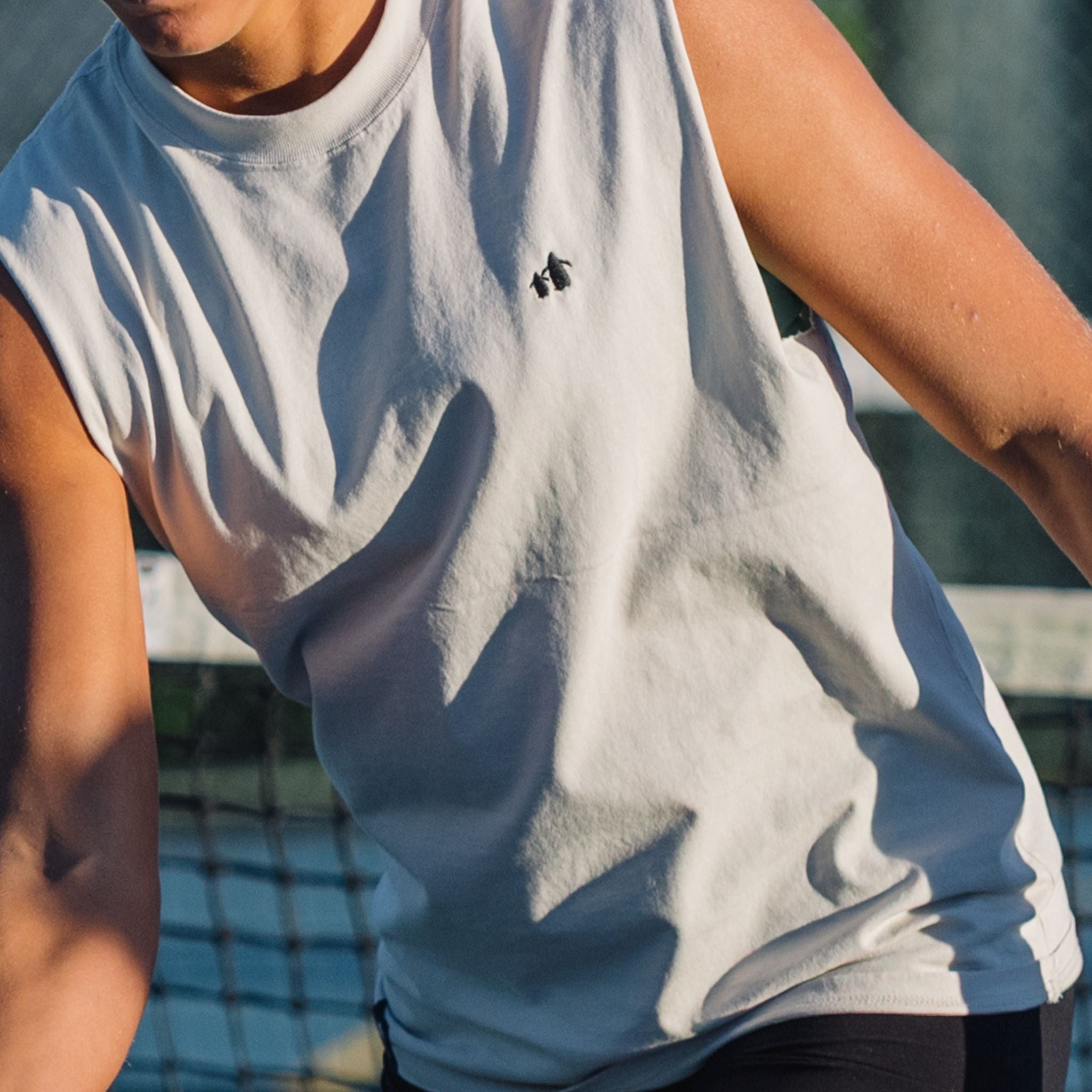 Person wearing a light bone sleeveless shirt with a small logo, standing outdoors.